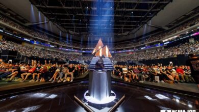 Imagem do troféu BLAST.tv Austin Major 2025 iluminado por luz branca e chamas do palco. Uma multidão lotada está ao fundo